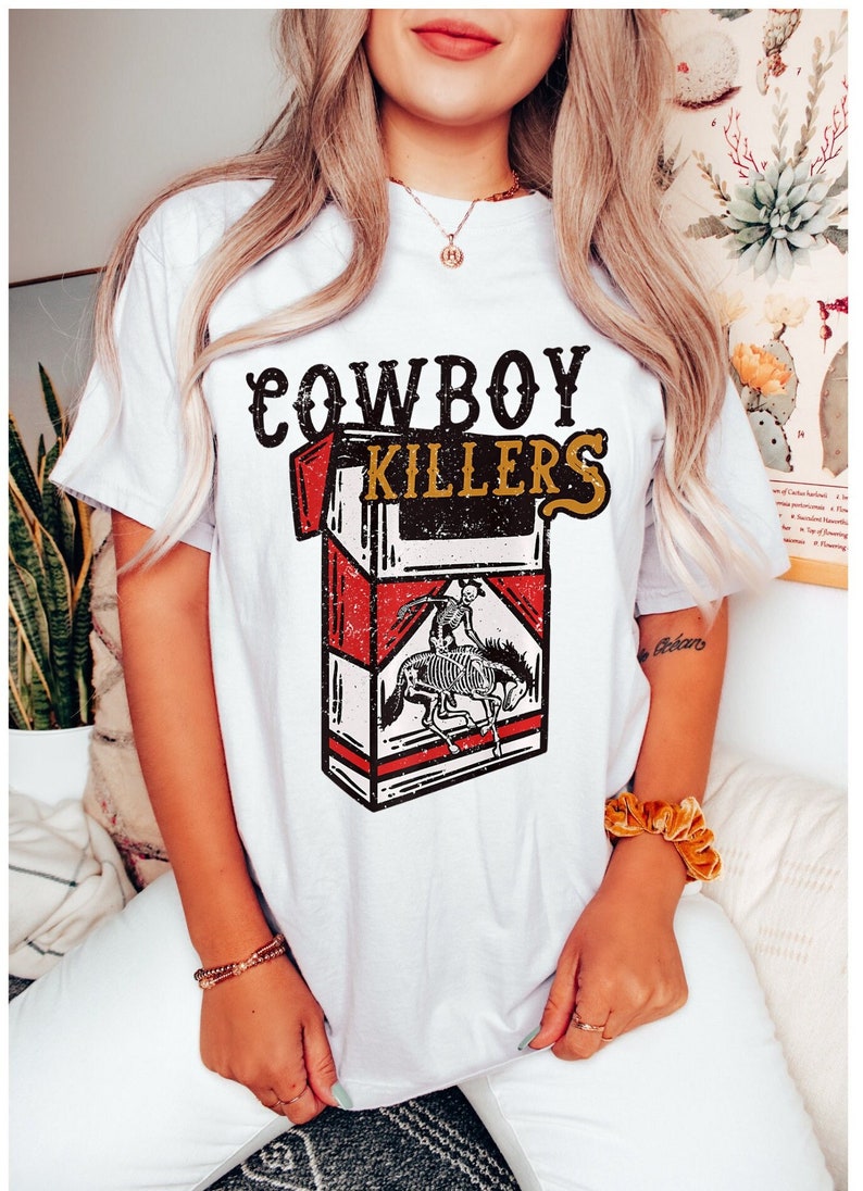 Peut inclure: T-shirt blanc avec un graphique vintage d'un paquet de cigarettes. Le paquet est rouge et blanc avec les mots "COWBOY KILLERS" imprim&eacute;s dessus. Un cow-boy squelette &agrave; cheval est repr&eacute;sent&eacute; sur le paquet.