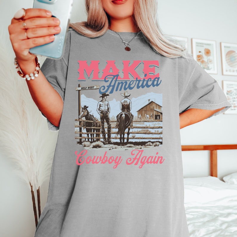 May include: Grey t-shirt featuring a graphic of two cowboys on horseback in front of a barn. The text "MAKE America Cowboy Again" is printed on the shirt.