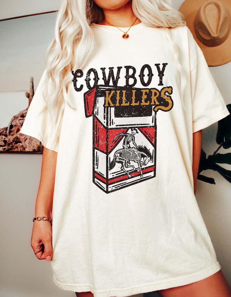 Peut inclure: T-shirt blanc avec un graphique repr&eacute;sentant un paquet de cigarettes avec un squelette &agrave; cheval. Le texte "COWBOY KILLERS" est imprim&eacute; au-dessus du graphique.
