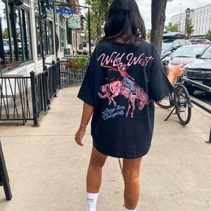 Puede incluir: Camiseta negra con un gr&aacute;fico de una vaquera montando a caballo, con las palabras "Wild West" y "Long live Cowgirls" en rosa y azul. La camiseta es de gran tama&ntilde;o y tiene mangas cortas.