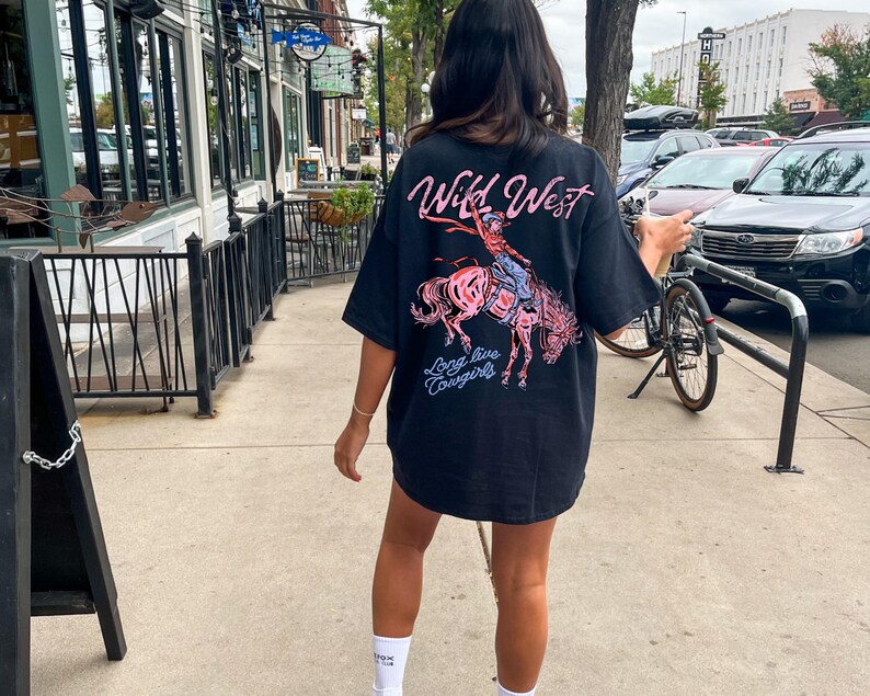 Puede incluir: Camiseta negra con un gr&aacute;fico de una vaquera montando a caballo, con las palabras "Wild West" y "Long live Cowgirls" en rosa y azul. La camiseta es de gran tama&ntilde;o y tiene mangas cortas.