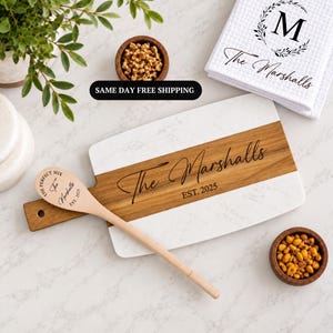May include: A rectangular serving board with white marble ends and a central wood panel engraved with The Marshalls EST. 2025, paired with a matching wooden spoon featuring the same text and a white waffle-weave towel with a monogrammed M.