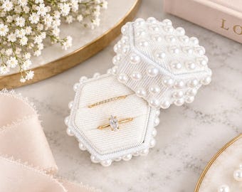 May include: A hexagonal white velvet ring box adorned with numerous small white pearls. The open box contains two gold rings, one with a marquise-cut stone and one with a thin band of small stones, resting in the plush white velvet interior.