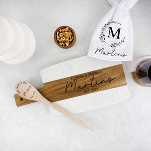 May include: A white marble and wood serving board with the text "Colleen & Michael Martins EST. 2024" engraved on it. A wooden spoon with the text "The Perfect Mix Martins EST. 2024" is on the board. A white dish towel with the letter "M" and the text "Martins" is in the background.