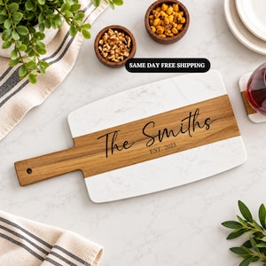 May include: A rectangular serving board featuring a white marble surface and a central horizontal strip of natural wood. The wood section is engraved with the text "The Smiths" and "EST. 2023" in a black script and sans-serif font.