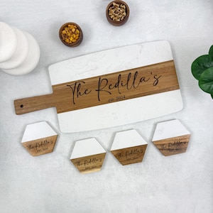 May include: A white marble and wood serving board with a wooden handle. The board has a white marble top and a wooden bottom with the text "The Redilla's est. 2024" engraved on it. There are four matching coasters with the same design and text.