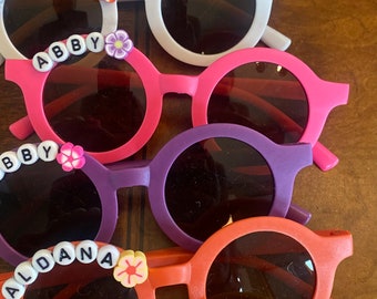 May include: A stack of colorful round sunglasses with different names spelled out in beads on the frames. The sunglasses are arranged from top to bottom with the names Mikaela, Greyson, Rolline, Abby, Abby, Aldana, Briana, and Brenna.