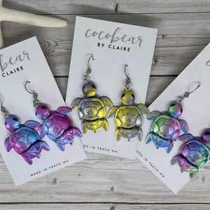 May include: Three pairs of turtle-shaped dangle earrings on white display cards labelled cocobear BY CLAIRE MADE IN PERTH WA. The earrings feature iridescent finishes in blue-purple, yellow-silver, and green-pink gradients with metal hooks.
