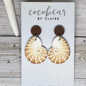 May include: A pair of earrings on a white display card labelled cocobear BY CLAIRE and MADE IN PERTH WA. Each earring features a small, dark brown circular wooden stud attached to a white and tan nautilus shell-shaped pendant with spiral details.