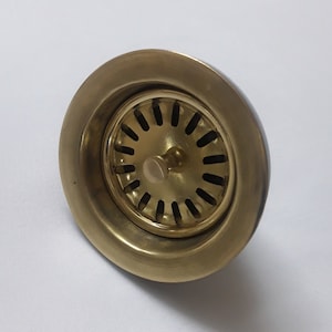 Könnte beinhalten: Ein goldfarbener Abflussstopfen aus Metall mit einem runden Design und einem kleinen Knopf in der Mitte.