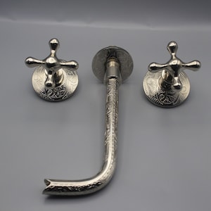 Puede incluir: Un grifo de baño plateado con tres manijas ornamentadas y un caño decorativo. Las manijas tienen un diseño en forma de cruz con detalles intrincados.