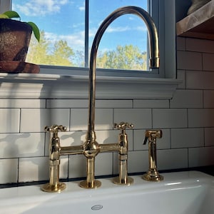 May include: A gold-toned kitchen faucet with a high-arc spout and two handles. The faucet is installed on a white tiled countertop with a white porcelain sink.