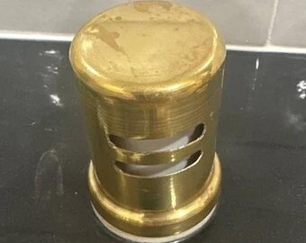 May include: A gold-colored metal drain stopper with a round top and two holes. The stopper is sitting on a black and white surface.