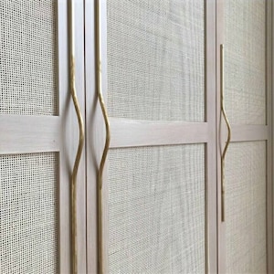 Op de afbeelding: Twee houten kastdeuren met geweven rieten panelen en gouden metalen handgrepen. De handgrepen hebben een uniek gebogen ontwerp.