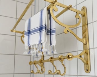 May include: A gold-coloured towel rail with a white and blue striped towel. The rail has decorative curved supports and hooks below. The towel has tassels. The rail is mounted on a white tiled wall.