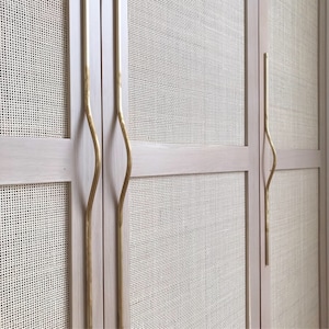 Op de afbeelding: Twee houten kastdeuren met geweven rieten panelen en gouden metalen handgrepen. De handgrepen hebben een uniek gebogen ontwerp.