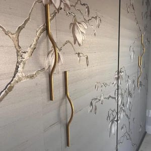 Puede incluir: Dos tiradores de puerta de armario de metal dorado con un diseño curvo están montados en una pared con un patrón de papel tapiz floral. El papel tapiz presenta un fondo marrón claro con un diseño floral blanco.