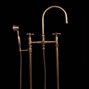 Könnte beinhalten: Eine freistehende goldfarbene Badewannenarmatur mit einer Handbrause. Die Armatur hat zwei Griffe und einen gebogenen Auslauf.