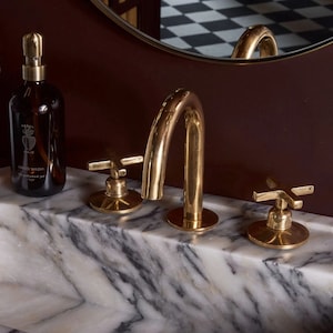 May include: A marble bathroom sink with three gold taps. The taps have a cross-shaped handle and a curved spout. The sink is partially filled with water.
