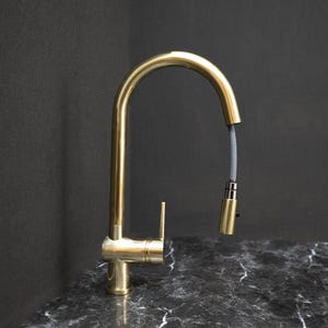 Pode incluir: Uma torneira de cozinha dourada com um bico de arco alto e um pulverizador retrátil. A torneira é feita de metal e tem um design elegante e moderno. O pulverizador retrátil está conectado a uma mangueira cinza. A torneira é montada em uma bancada preta e cinza.