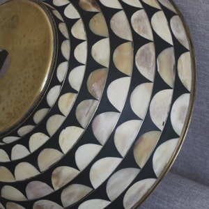 Puede incluir: Un objeto decorativo circular con una parte superior de color lat&oacute;n y una superficie estampada. La superficie presenta un dise&ntilde;o repetitivo de formas semicirculares de color crema sobre un fondo negro, enmarcado por un borde de color lat&oacute;n.