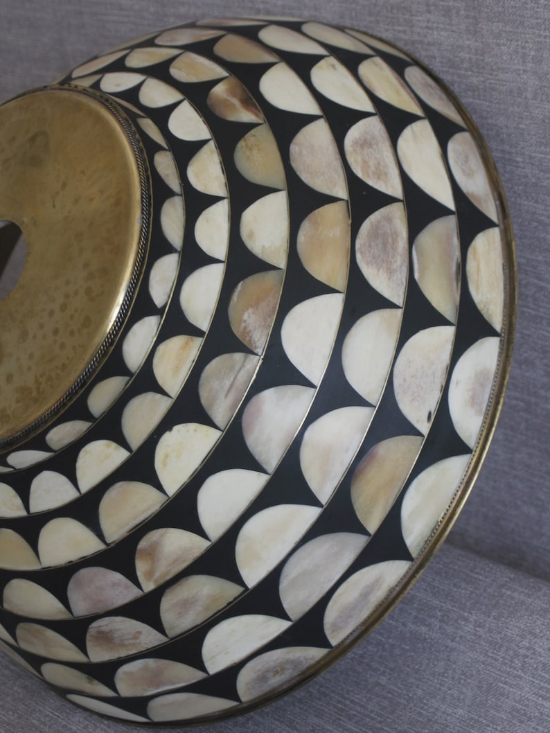 Puede incluir: Un objeto decorativo circular con una parte superior de color lat&oacute;n y una superficie estampada. La superficie presenta un dise&ntilde;o repetitivo de formas semicirculares de color crema sobre un fondo negro, enmarcado por un borde de color lat&oacute;n.