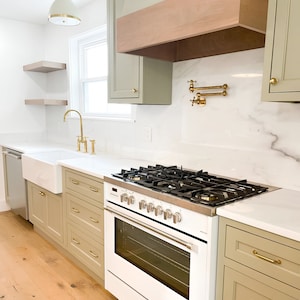 May include: A white kitchen with light green cabinets, a farmhouse sink, and a stainless steel range with a white oven. The countertop is white marble with gold hardware.