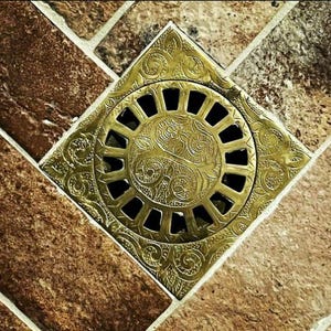 Könnte beinhalten: Ein quadratischer Abflussdeckel in Gold mit einem filigranen Blumenmuster. Der Abflussdeckel ist in einen Fliesenboden eingelassen.
