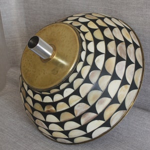 Puede incluir: Pantalla de l&aacute;mpara decorativa con parte superior de lat&oacute;n y dise&ntilde;o estampado. La pantalla presenta un patr&oacute;n repetitivo de semic&iacute;rculos blanquecinos y negros. Un tubo de metal plateado se extiende desde la parte superior de lat&oacute;n.