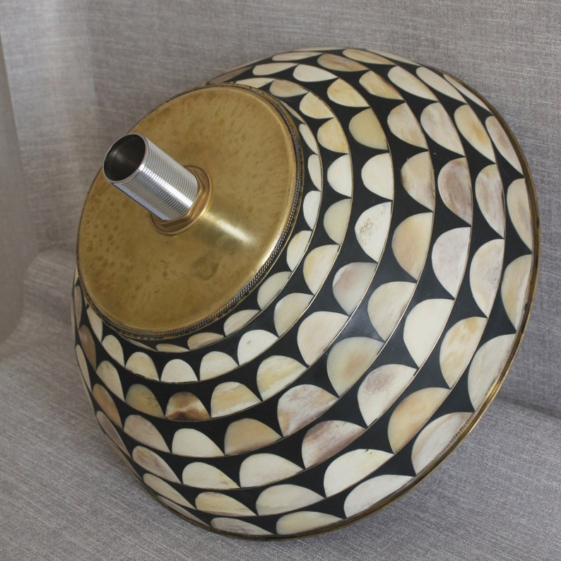 Puede incluir: Pantalla de l&aacute;mpara decorativa con parte superior de lat&oacute;n y dise&ntilde;o estampado. La pantalla presenta un patr&oacute;n repetitivo de semic&iacute;rculos blanquecinos y negros. Un tubo de metal plateado se extiende desde la parte superior de lat&oacute;n.