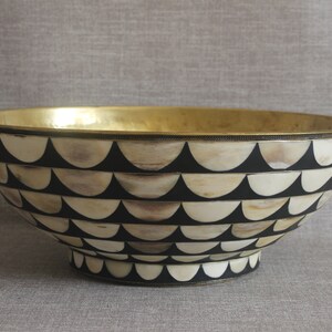 Puede incluir: Un cuenco decorativo con interior dorado y exterior estampado. El exterior presenta un dise&ntilde;o repetitivo de semic&iacute;rculos negros y crema. El cuenco tiene una forma amplia y abierta y una peque&ntilde;a base elevada.