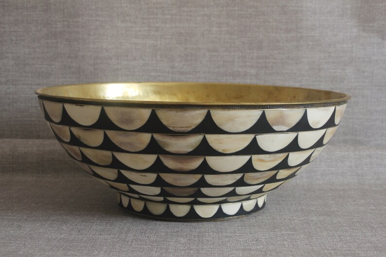 Puede incluir: Un cuenco decorativo con interior dorado y exterior estampado. El exterior presenta un dise&ntilde;o repetitivo de semic&iacute;rculos negros y crema. El cuenco tiene una forma amplia y abierta y una peque&ntilde;a base elevada.
