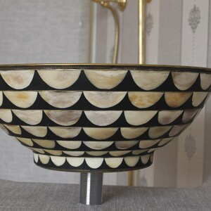 Puede incluir: Un lavabo decorativo con un dise&ntilde;o &uacute;nico. La base presenta un fondo negro con un patr&oacute;n de formas semicirculares de color crema. El borde y la base est&aacute;n adornados con un metal dorado. El lavabo est&aacute; montado sobre una tuber&iacute;a cromada.