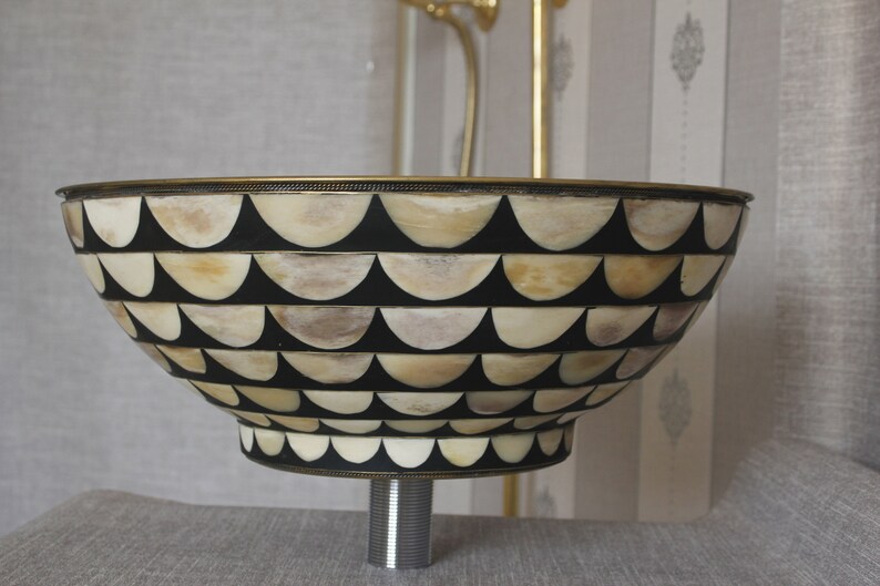 Puede incluir: Un lavabo decorativo con un dise&ntilde;o &uacute;nico. La base presenta un fondo negro con un patr&oacute;n de formas semicirculares de color crema. El borde y la base est&aacute;n adornados con un metal dorado. El lavabo est&aacute; montado sobre una tuber&iacute;a cromada.