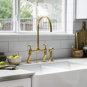 May include: A white double-bowl kitchen sink with a gold-toned bridge tap. The tap has two handles and a separate sprayer. The sink is set against a white tiled backsplash.