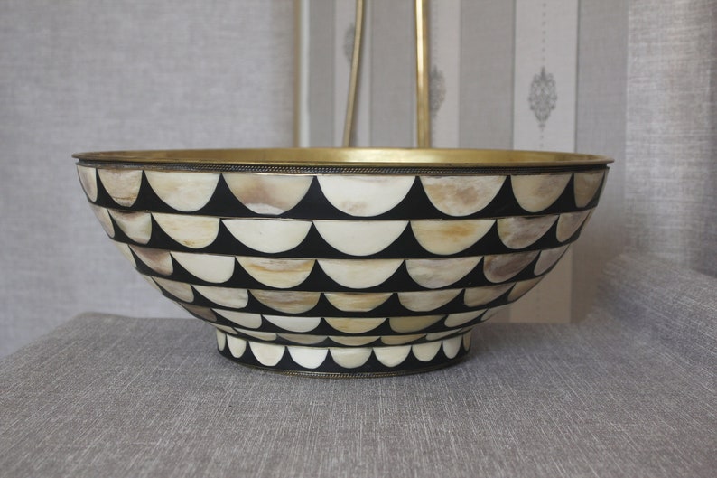 Puede incluir: Cuenco decorativo con borde de lat&oacute;n y un patr&oacute;n de semic&iacute;rculos negros y crema alternados. El cuenco tiene un dise&ntilde;o amplio y abierto y una base decorativa. Probablemente se utiliza para exhibici&oacute;n o servicio.