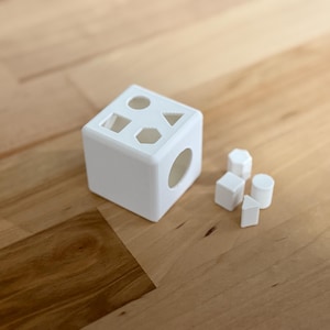 May include: White wooden shape sorter toy with five different shaped blocks. The blocks are a circle, square, triangle, pentagon, and a rectangle.