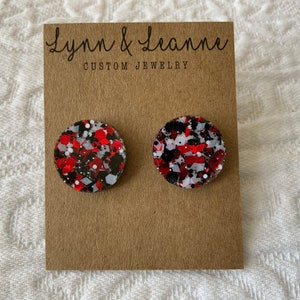 May include: A pair of round stud earrings with a glittery design in red, black, white, and gray. The earrings are displayed on a brown card with the text "Lynn & Leanne CUSTOM JEWELRY".