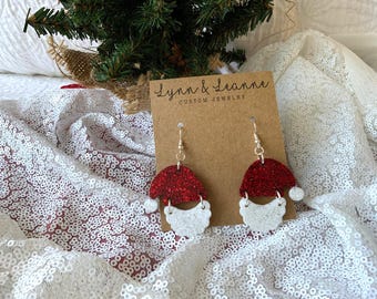 May include: A pair of festive Santa hat earrings with red glitter tops and white beard accents. The earrings are displayed on a brown card with the text "Lynn & Leanne CUSTOM JEWELRY". The background features a white sequined fabric and a small Christmas tree.