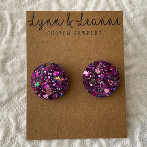May include: A pair of round, purple glitter earrings. The earrings are on a brown card with the text "Lynn & Leanne CUSTOM JEWELRY".