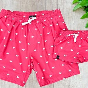 May include: Two pairs of red swimming trunks with a white whale pattern. The larger pair has a drawstring waist and the smaller pair has an elastic waist.