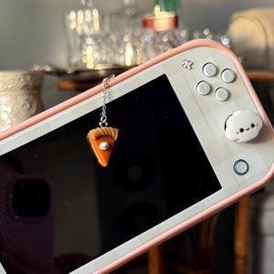 May include: A white and pink handheld gaming console with a pie slice charm. The console has white buttons and a black screen. The charm is a small, orange pie slice with a silver chain.