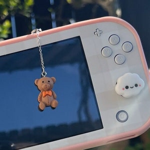 Könnte beinhalten: Eine rosa Nintendo Switch Lite mit einem weißen, wolkenförmigen Knopf auf der linken Seite und einem weißen, wolkenförmigen Knopf mit einem Smiley auf der rechten Seite. Ein brauner Teddybär-Anhänger hängt an der Oberseite des Geräts.