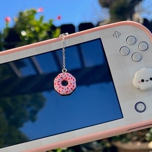 May include: A pink and white Nintendo Switch Lite handheld gaming console with a pink donut charm hanging from the top screen. The console has a white cloud-shaped button on the left side and a white cloud-shaped button on the right side.