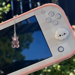 May include: A pink Nintendo Switch Lite with a white and pink checkered case. The console has a cloud-shaped button with a smiley face on the left side and a cloud-shaped button with a smiley face on the right side. A pink gummy bear charm hangs from the top of the console.