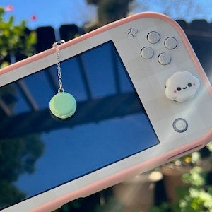 Puede incluir: Una Nintendo Switch Lite rosa con un amuleto de macaron verde colgando de la parte superior. La consola tiene botones blancos y una cubierta de botón en forma de nube blanca.