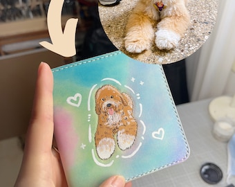 May include: A small, light blue leather wallet with a hand-painted illustration of a brown and white dog. The dog is sitting with its paws in the air and has a pink tongue sticking out. The background of the wallet is a pastel rainbow.