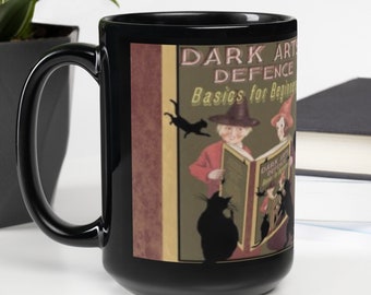 May include: A black ceramic mug featuring a graphic print of a book titled Dark Arts Defence Basics for Beginners. The illustration depicts two figures in pointed hats reading a book, surrounded by black cat silhouettes on a muted green background.