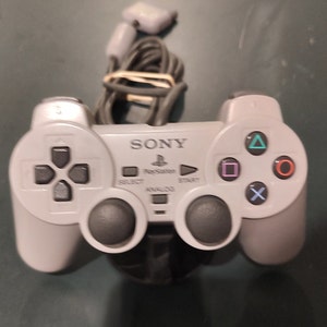 Pode incluir: Um controlador PlayStation Sony cinza com um joystick analógico preto e botões frontais. O controlador tem um logotipo PlayStation preto e branco e a palavra "SONY" impressa na frente.