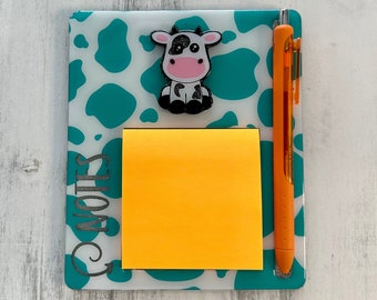 May include: A white and teal memo board with a cow design. The board has a yellow sticky note and an orange pen.